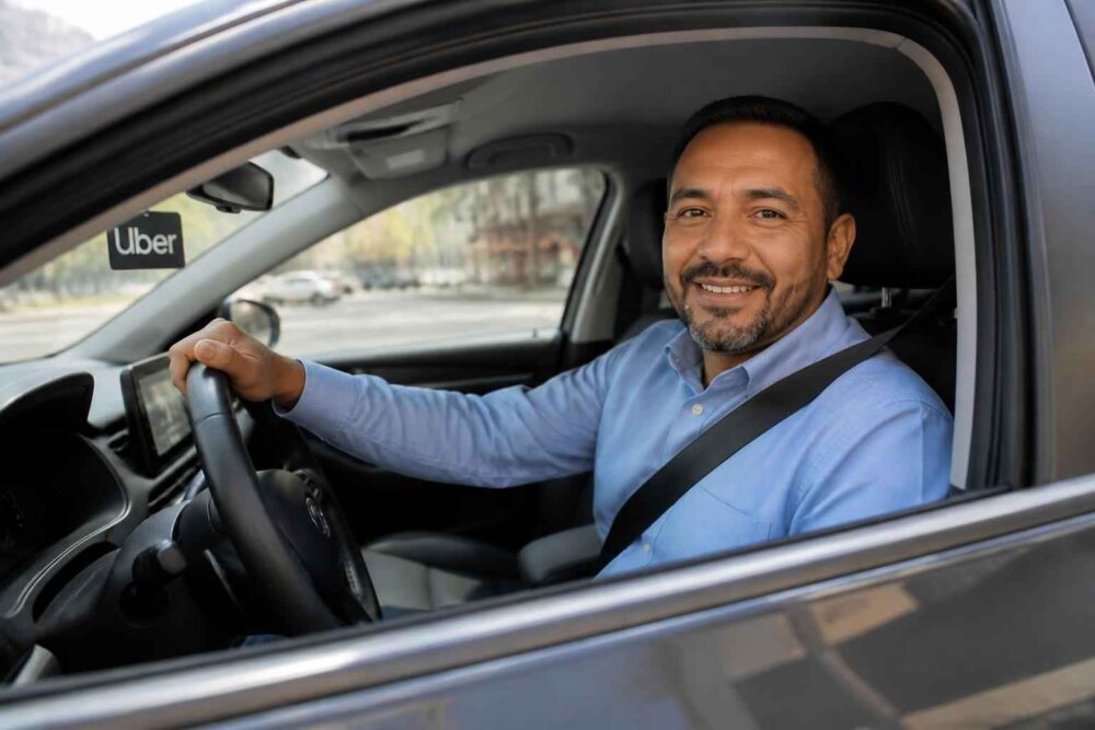 Uśmiechnięty kierowca Ubera siedzi za kierownicą samochodu z zapiętymi pasami bezpieczeństwa