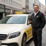 Taksówkarz w czarnym uniformie stoi obok białego mercedesa z oznaczeniem TAXI na miejskiej ulicy